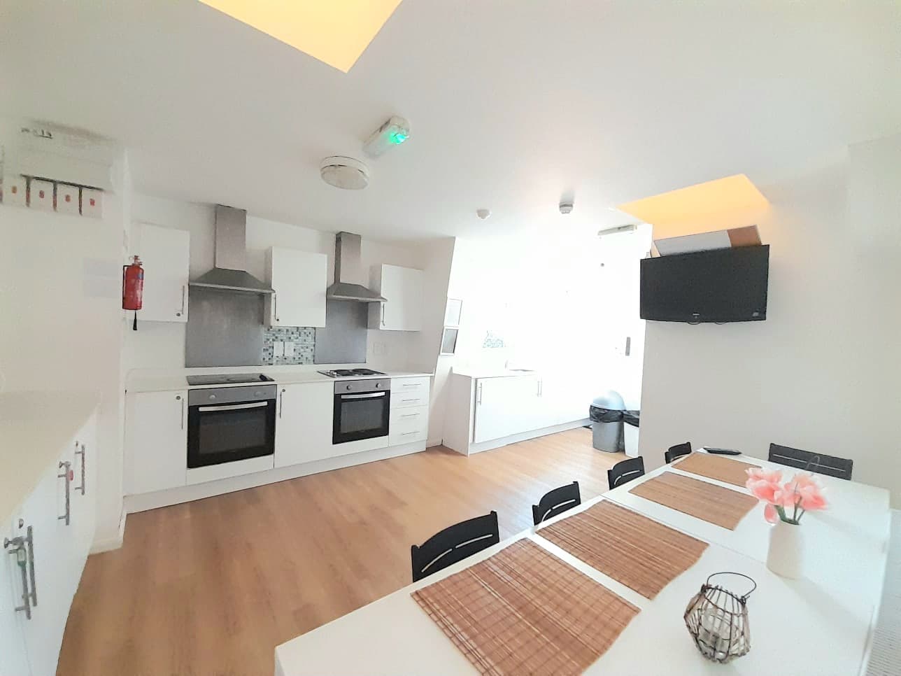 Modern white kitchen featuring dual ovens, wooden flooring, and a dining table with pink flowers.