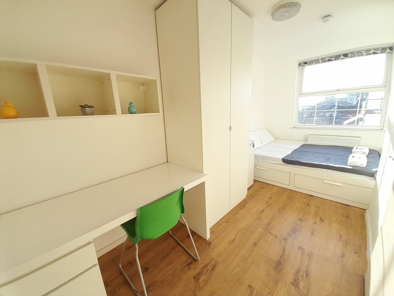 Bright, compact bedroom with a white desk, green chair, tall wardrobe, and single bed.
