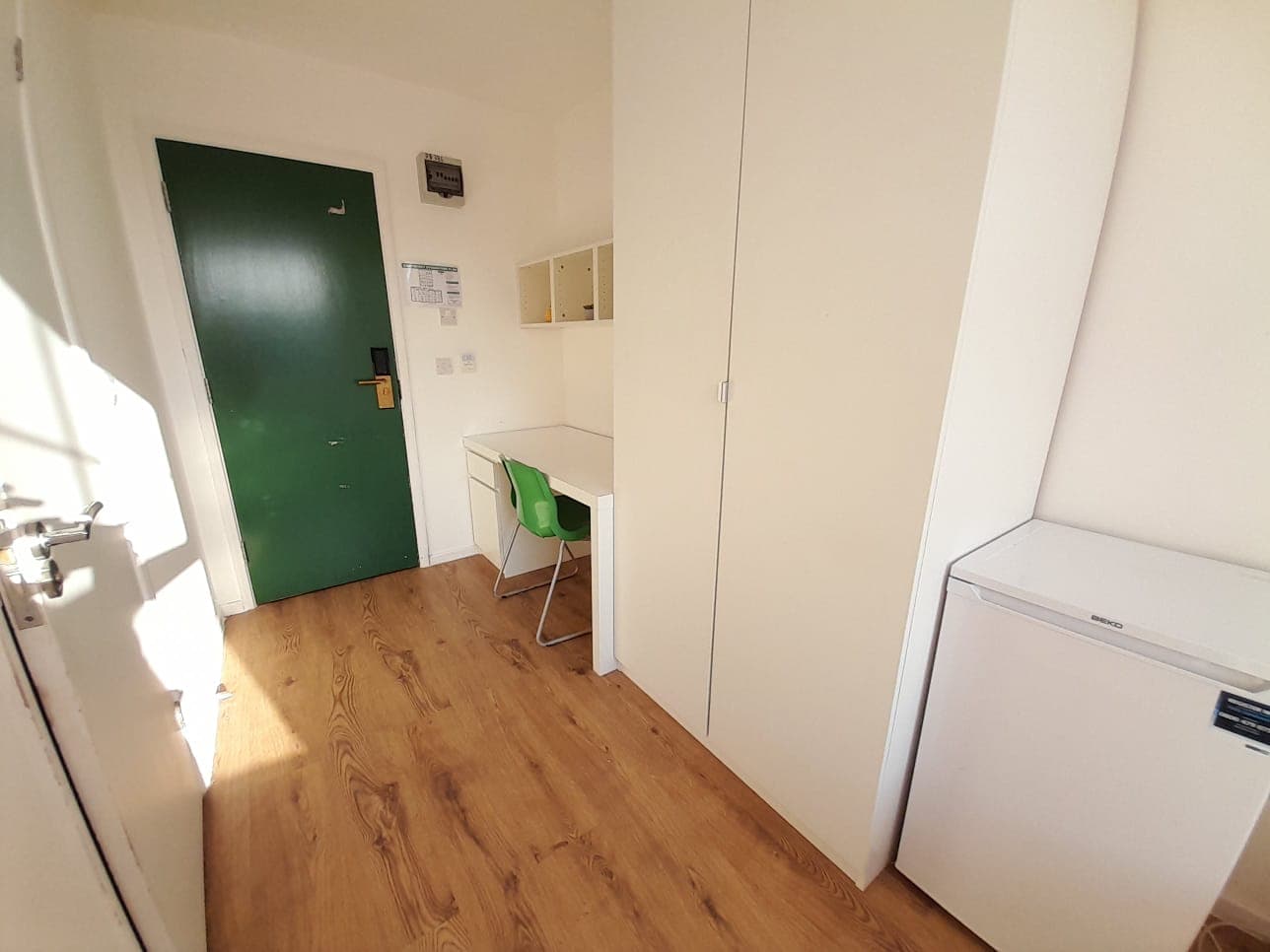 Compact living space with a green door, white desk, green chair, wardrobe, and mini-fridge.